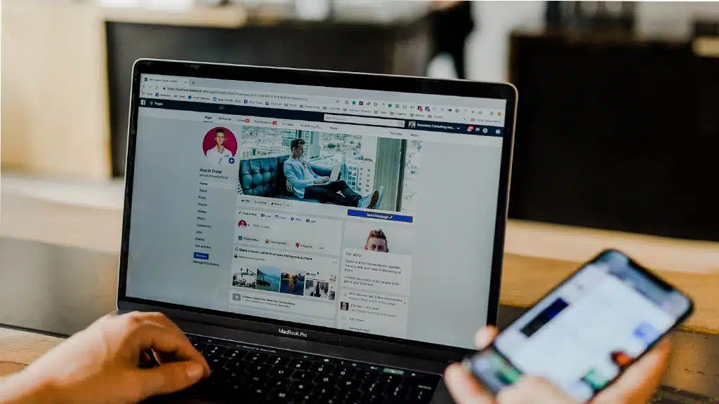 Person using a laptop displaying a Facebook business page, while holding a smartphone, emphasizing social media engagement.