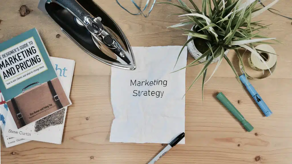 A desk with books on marketing and pricing, a plant, and a piece of paper labeled 'Marketing Strategy,' alongside markers and an iron.