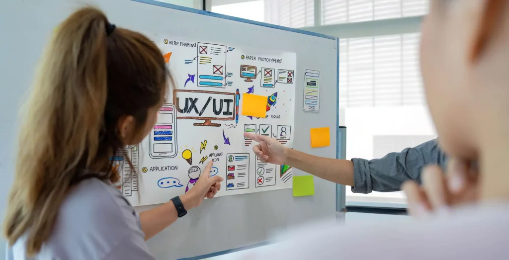 A woman points at a whiteboard adorned with various sticky notes, highlighting key ideas during a collaborative meeting