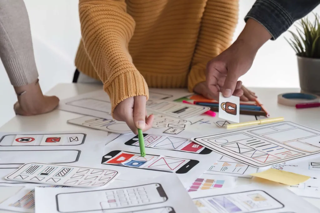 Professionals brainstorming and creating a mobile app design, surrounded by digital devices and design materials in an office setting.