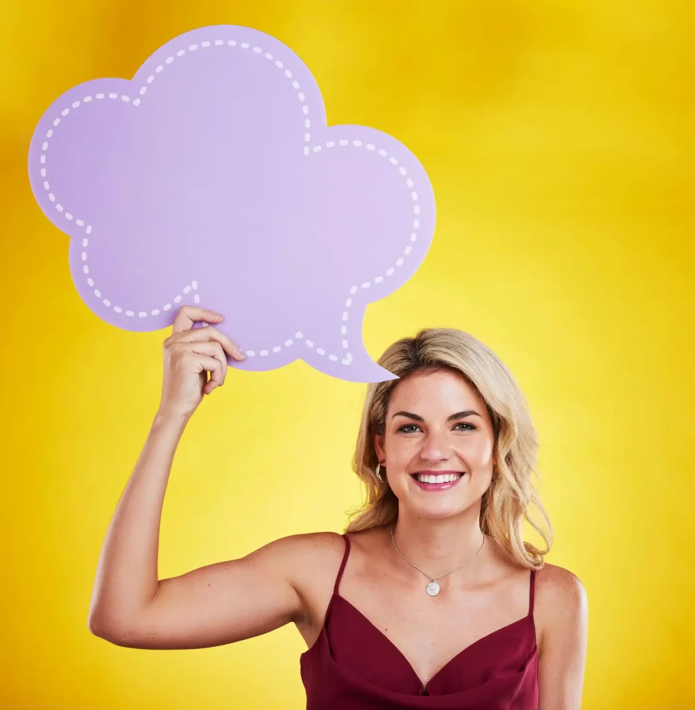 A woman with blonde hair in a burgundy top holds a large, purple, blank speech bubble above her head against a yellow background.