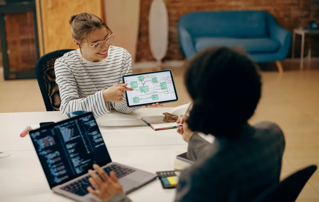 Two women collaborating at a table, with a laptop and a tablet in front of them.